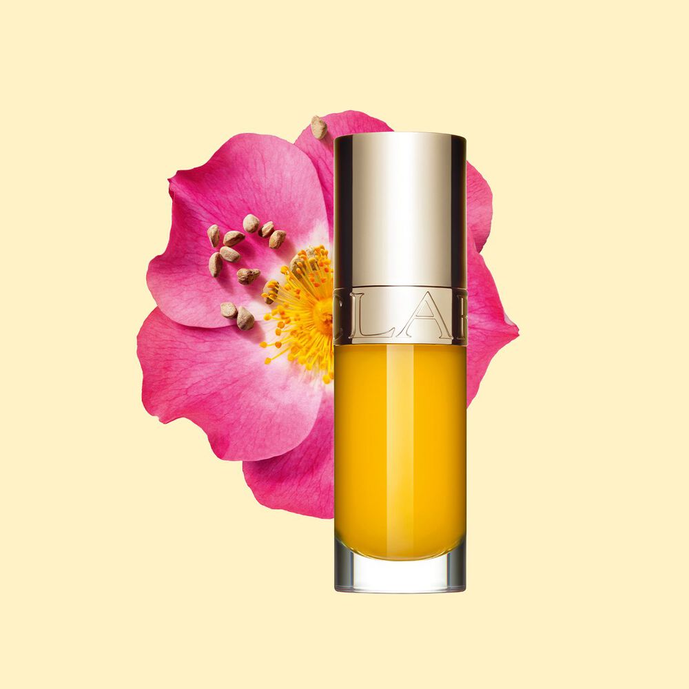 Foto del aceite labial amarillo de Clarins delante de una flor de rosa mosqueta con un fondo blanco