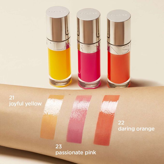 Presentación de la gama de aceites labiales Clarins en amarillo, rosa y naranja, con paleta de colores en un brazo de mujer